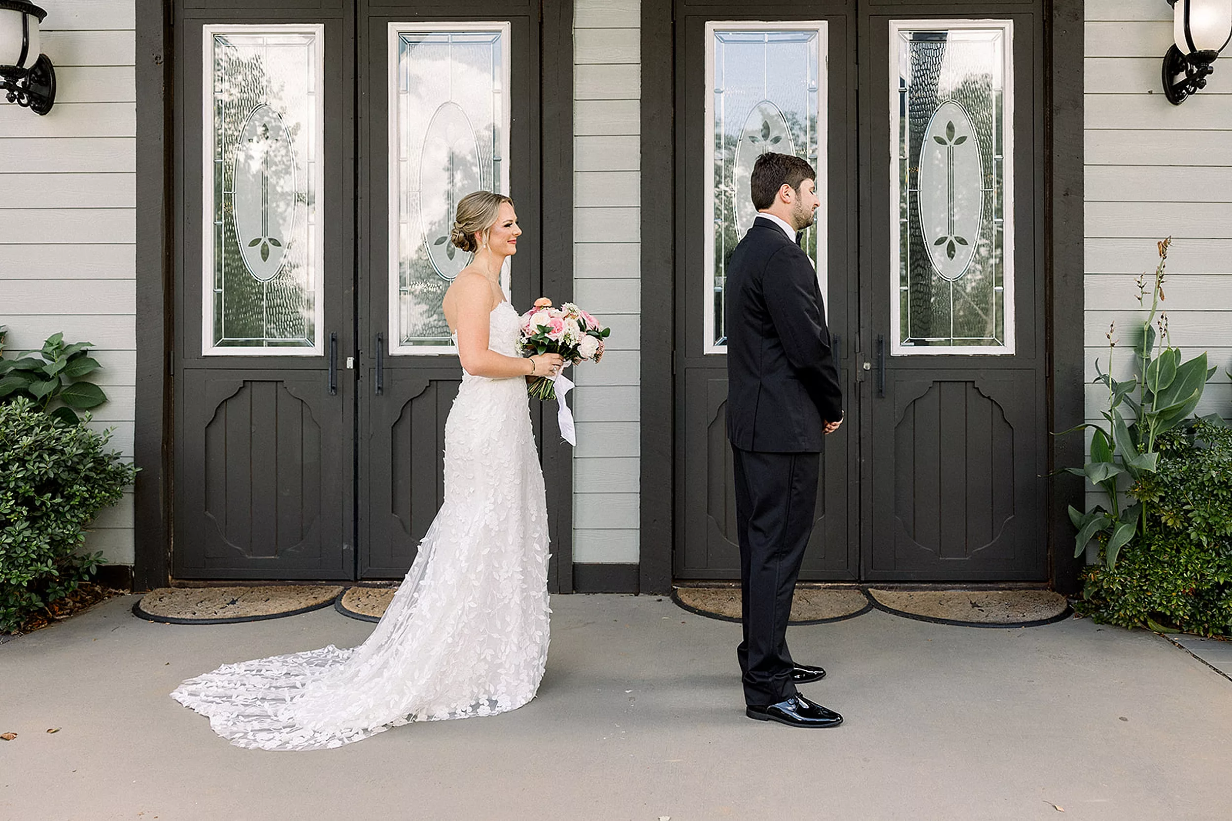 A groom stands with hands crossed in front of him with his back to his bride as she approaches for a first look on a porch