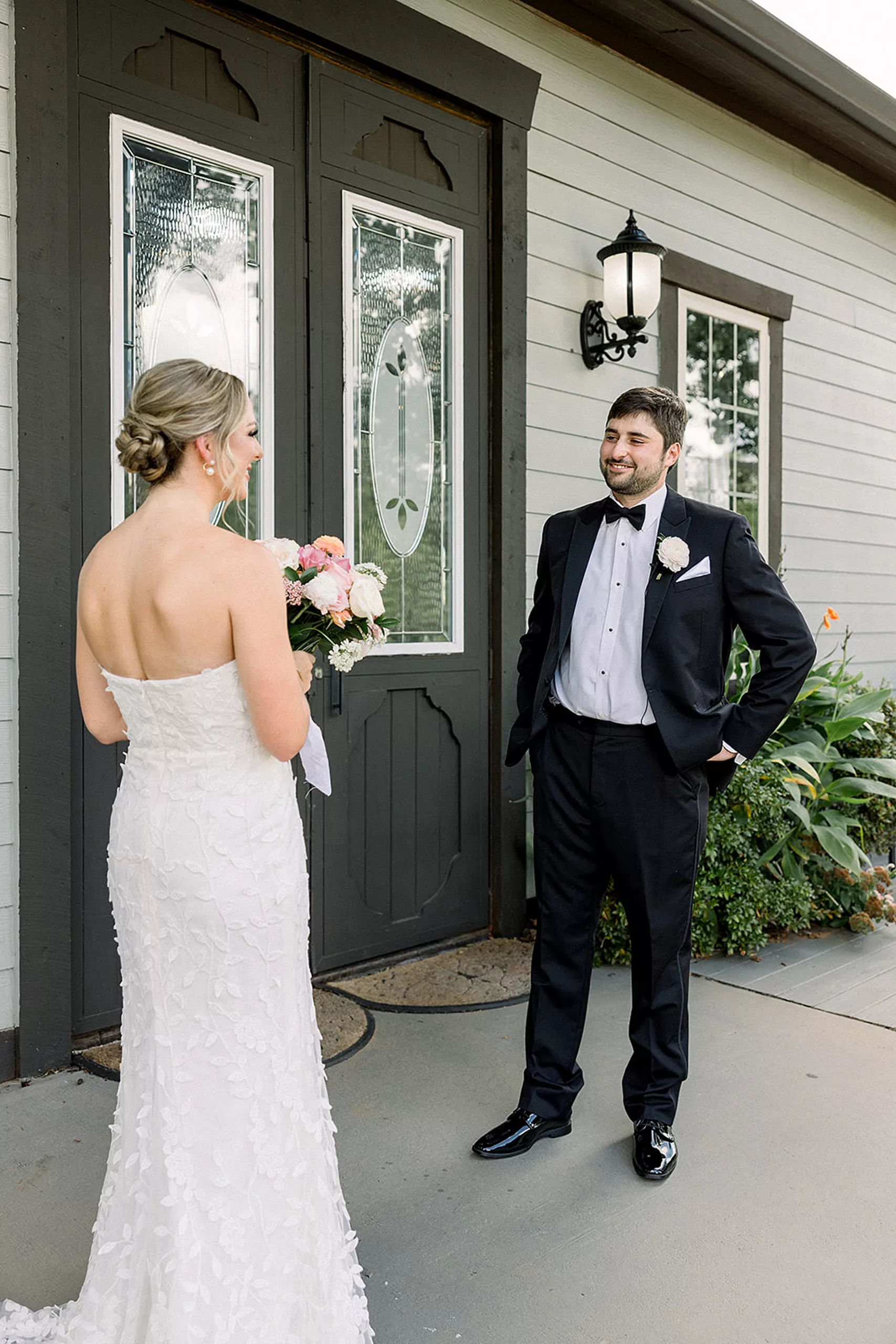 A groom stands with hands in his black suit pockets as he smiles and sees his bride in her dress for the first time