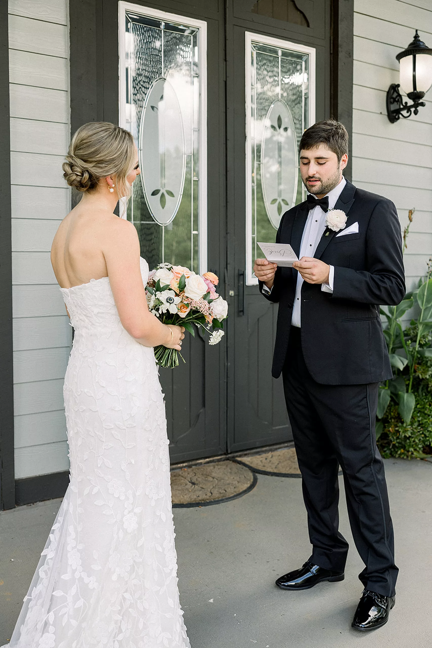 A groom in a black suit reads a letter with his bride while standing on a porch