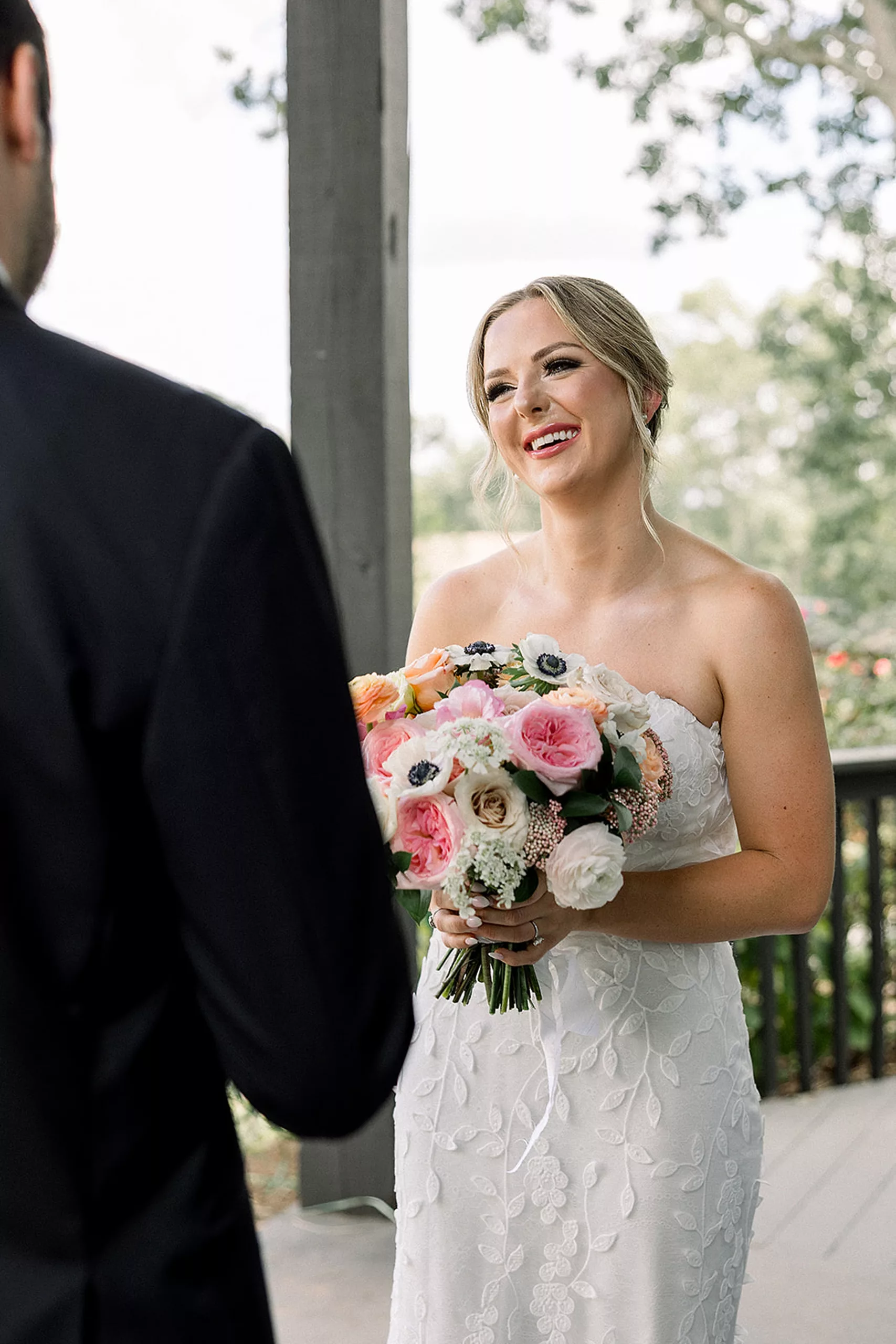 A bride smiles at her groom while holding her pink and white bouquet on a porch