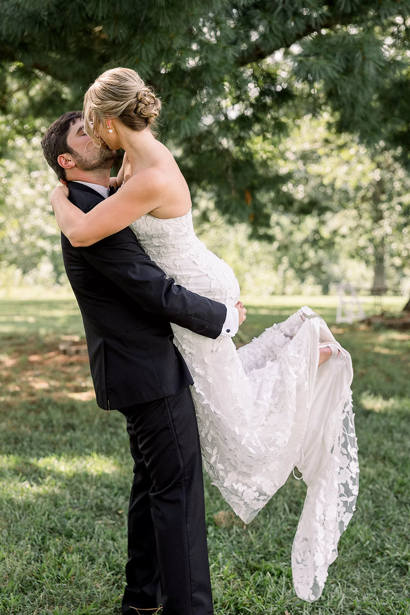 A groom lifts and kisses his bride in a field under trees
