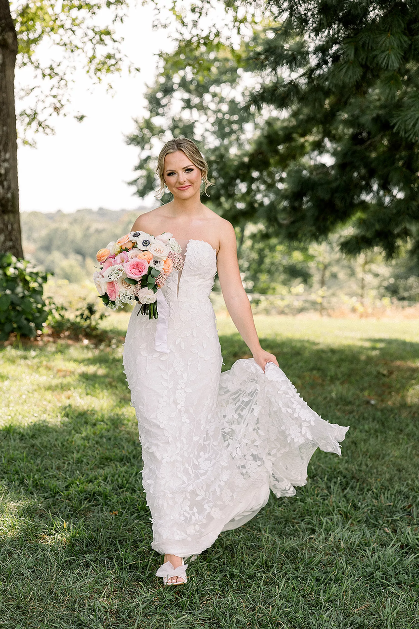 A bride walks through a grassy field under trees holding her train and her colorful bouquet in a lace dress