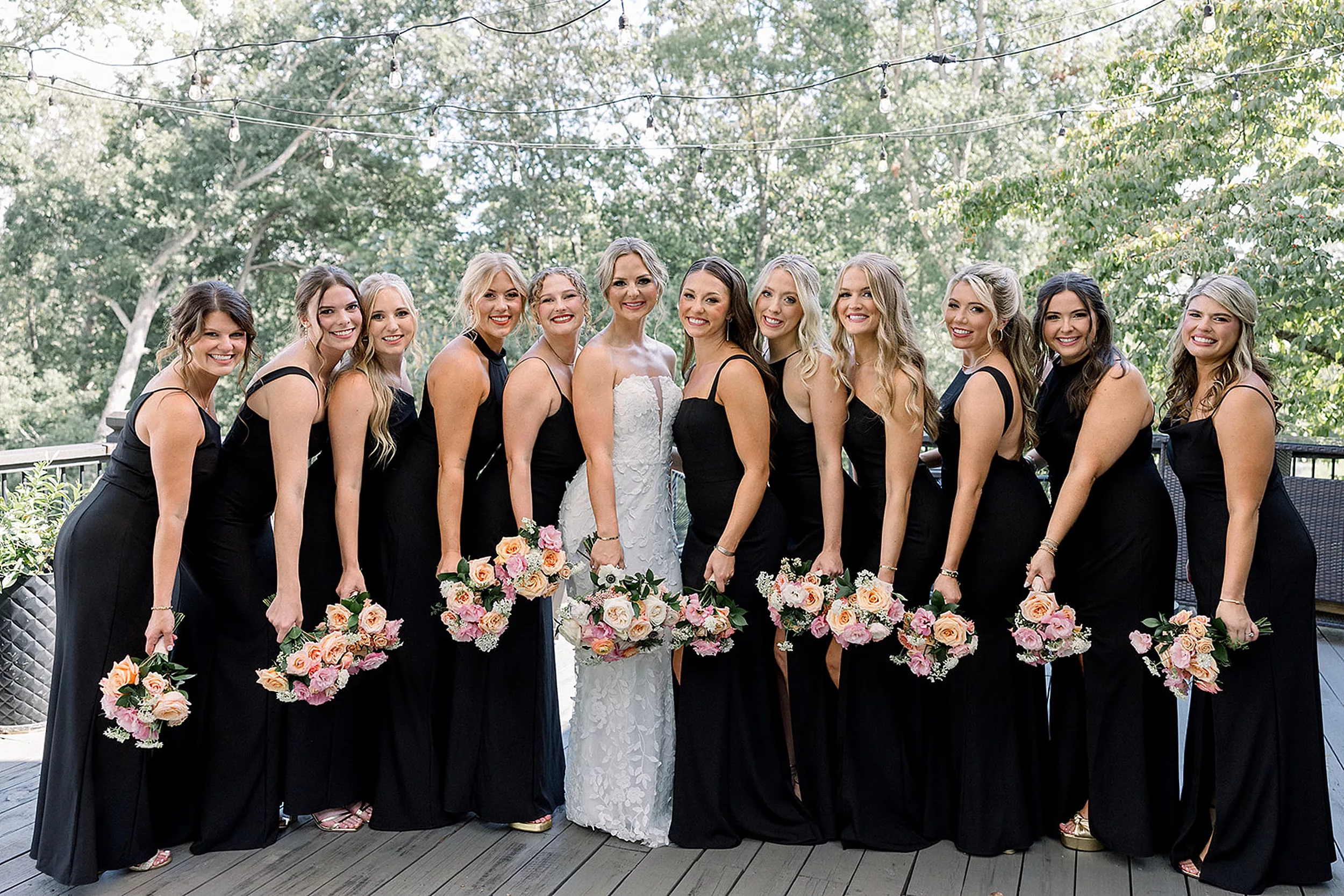 A bride stands on a porch holding her flowers with her large bridal party leaning in holding theirs in black dresses at White Oaks Vineyard