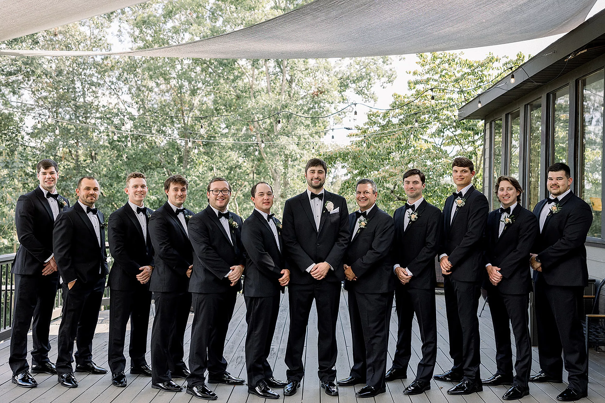 A groom in a black suit stands with hands crossed in front of him on a porch with his large wedding party in all black suits at White Oaks Vineyard
