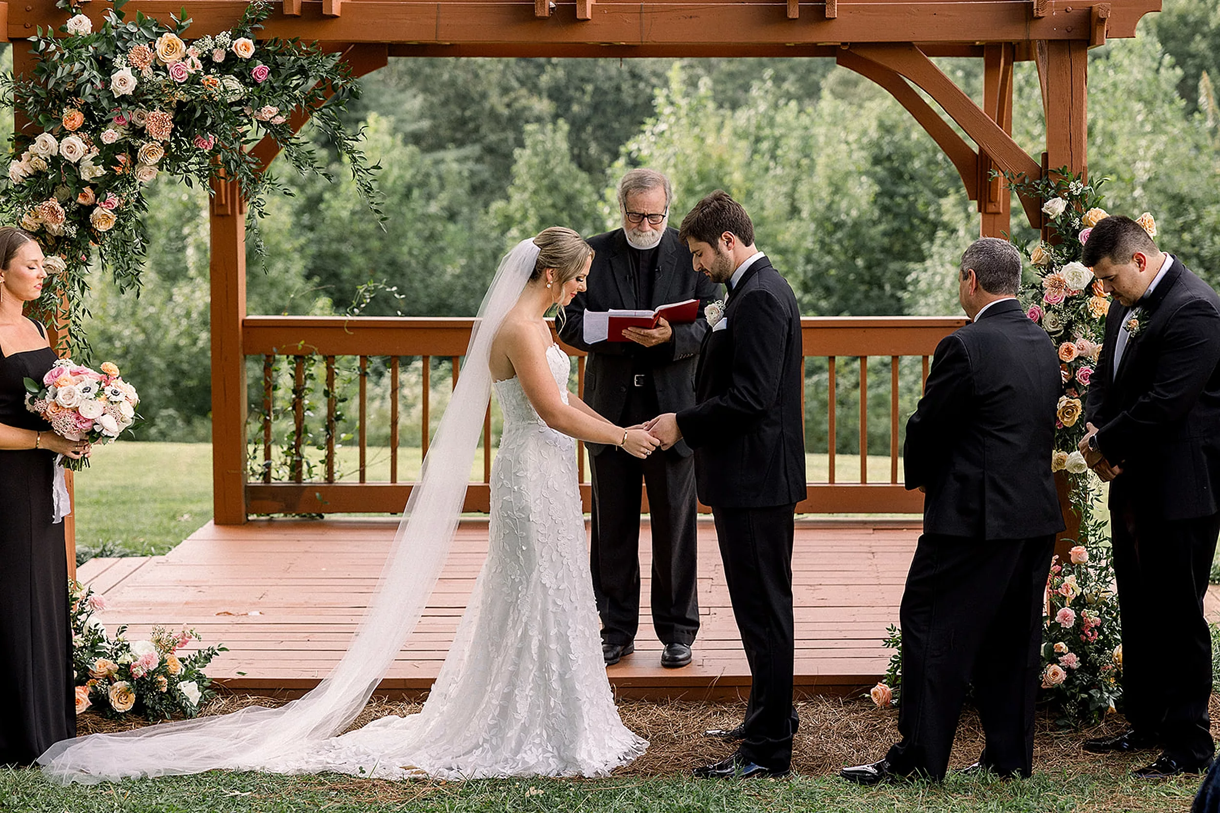 Newlyweds look down at their hands together during their ceremony under a brown pergola at a White Oaks Vineyard wedding ceremony