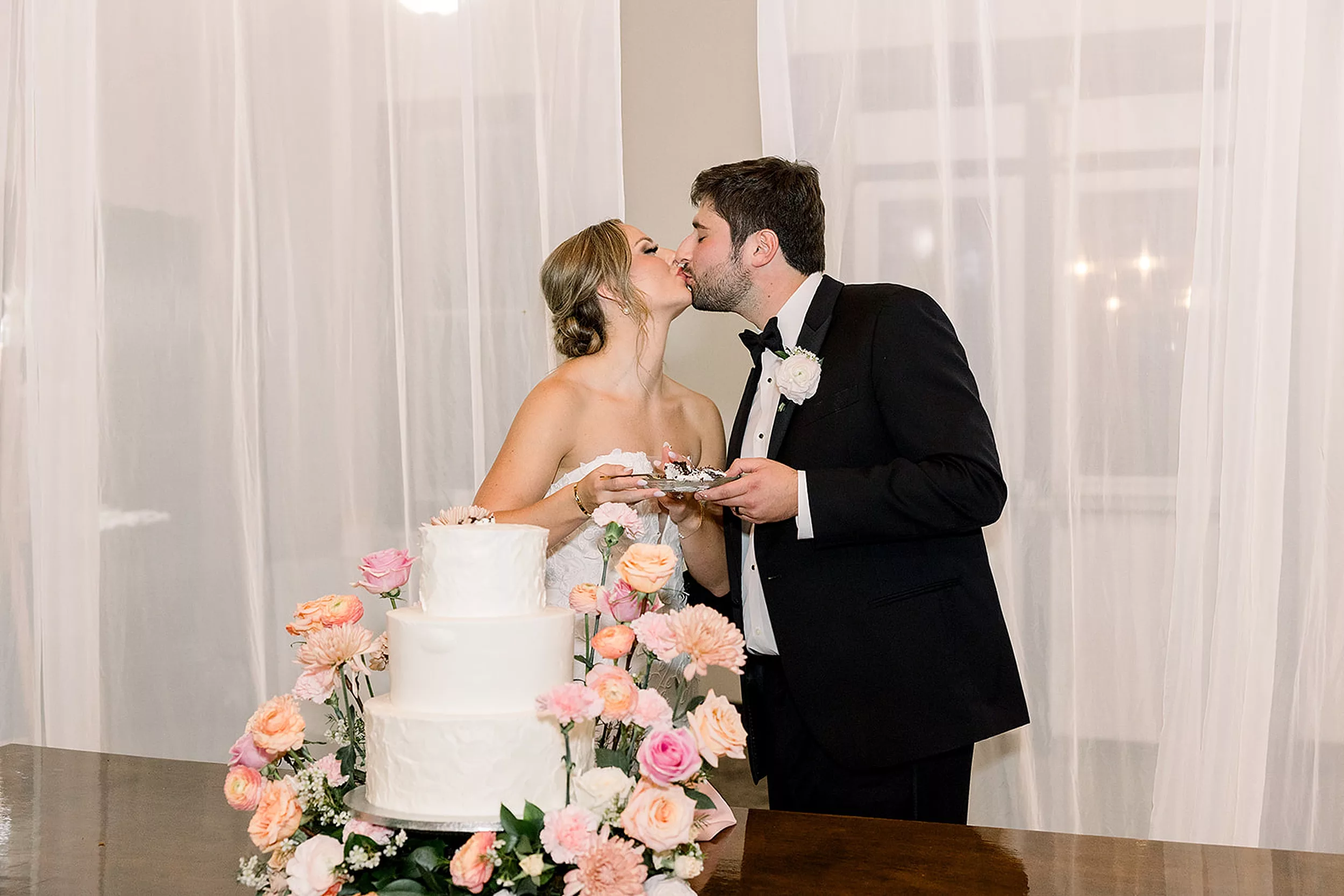 Newlyweds kiss after cutting their three tier cake in a black tuxedo and a lace dress at a White Oaks Vineyard wedding reception