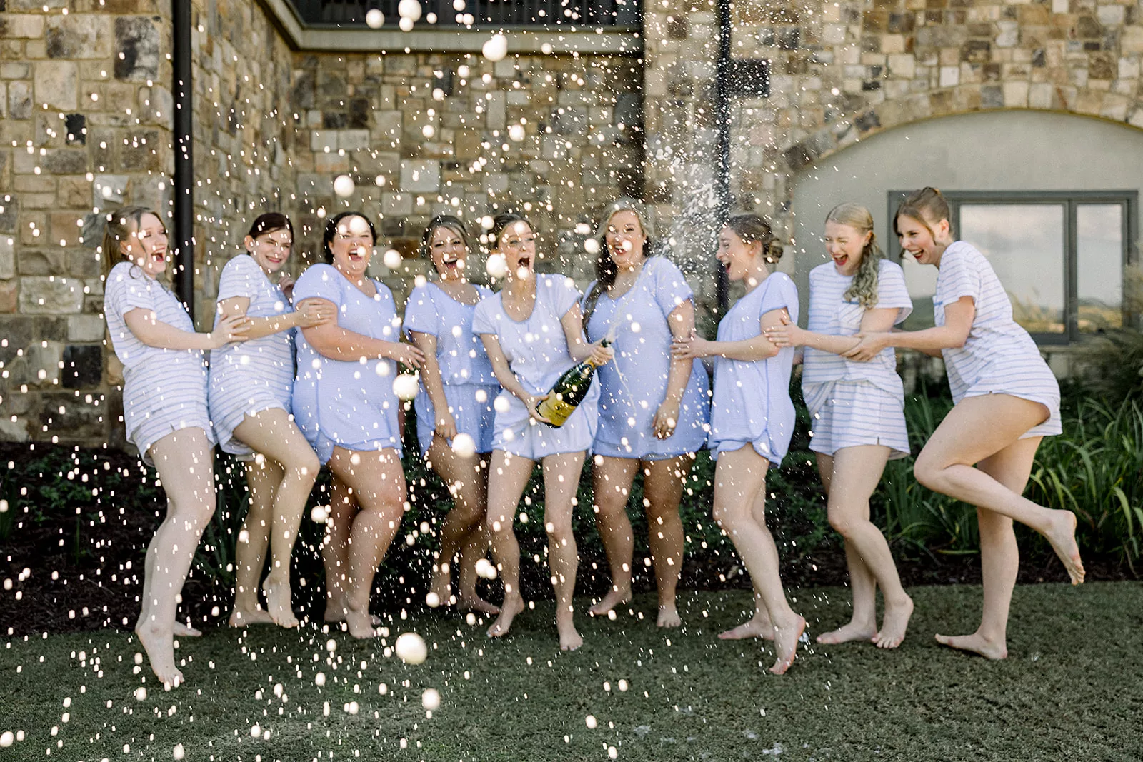 A bride pops a bottle of a champagne while wearing blue pajamas with her bridesmaids
