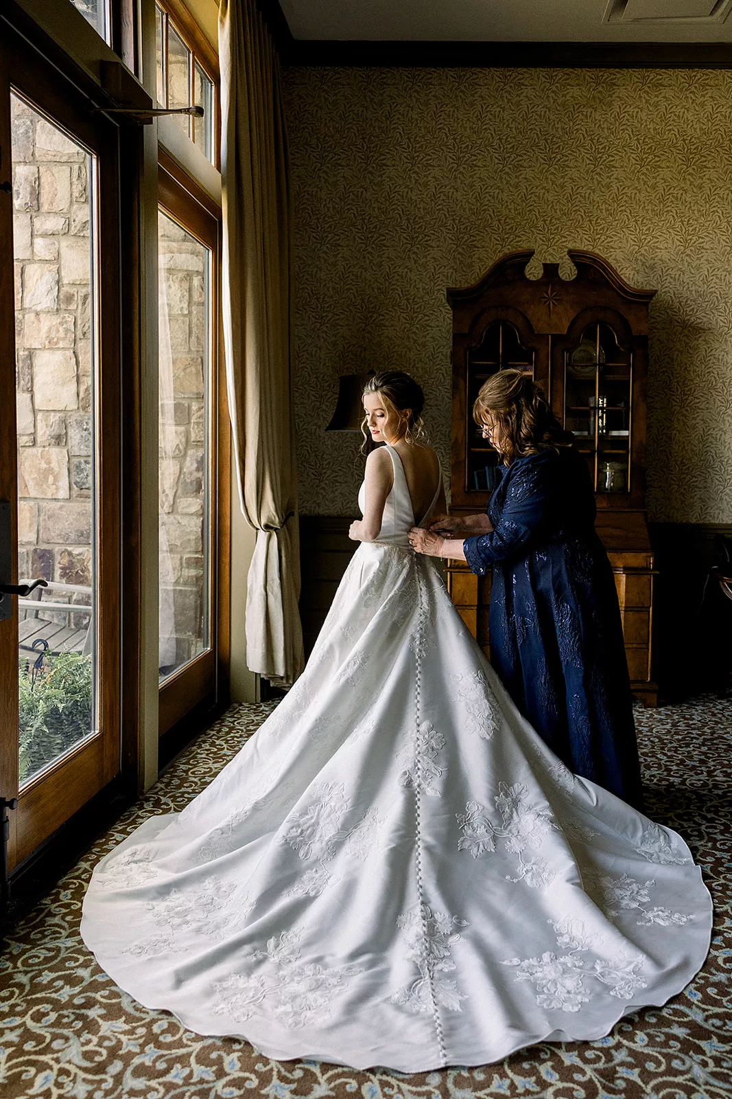 A bride stands in an antique room getting buttoned into her large dress by her mother