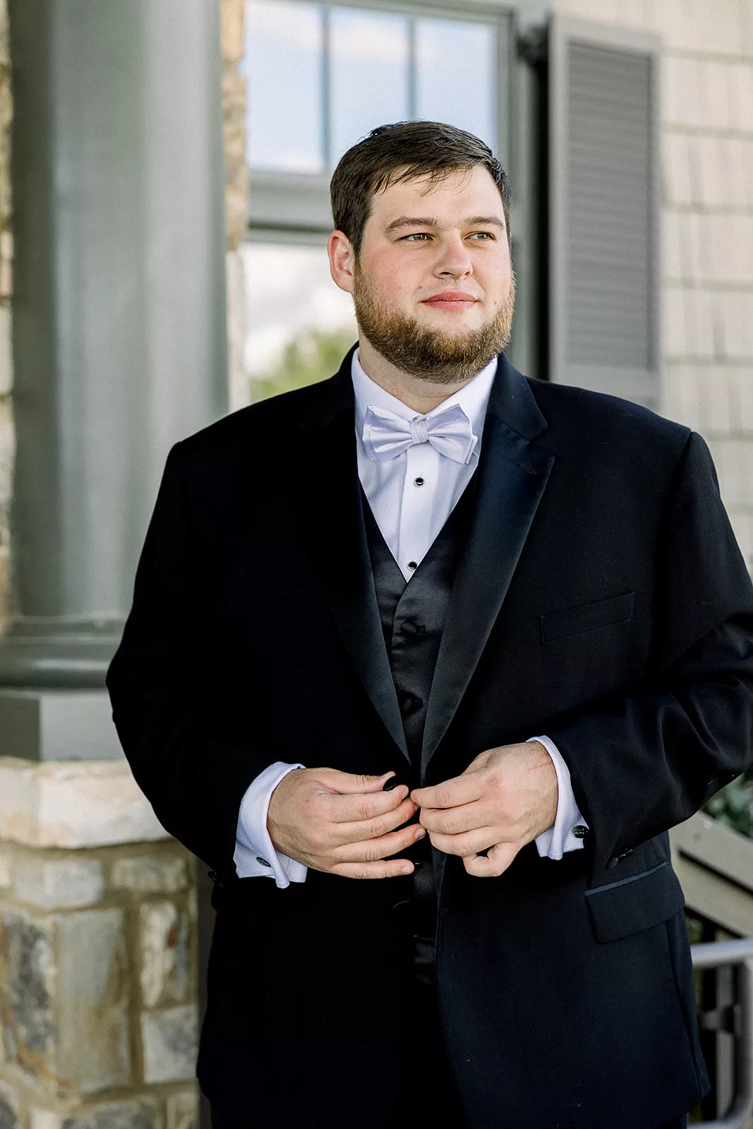A groom stands outside a stone building buttoning his black tuxedo jacket
