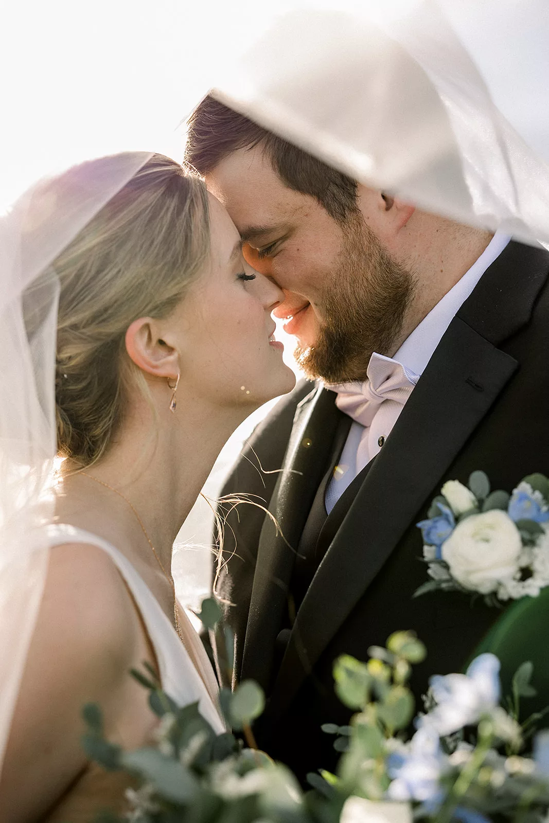 Newlyweds lean in for a kiss while hiding under a veil at sunset