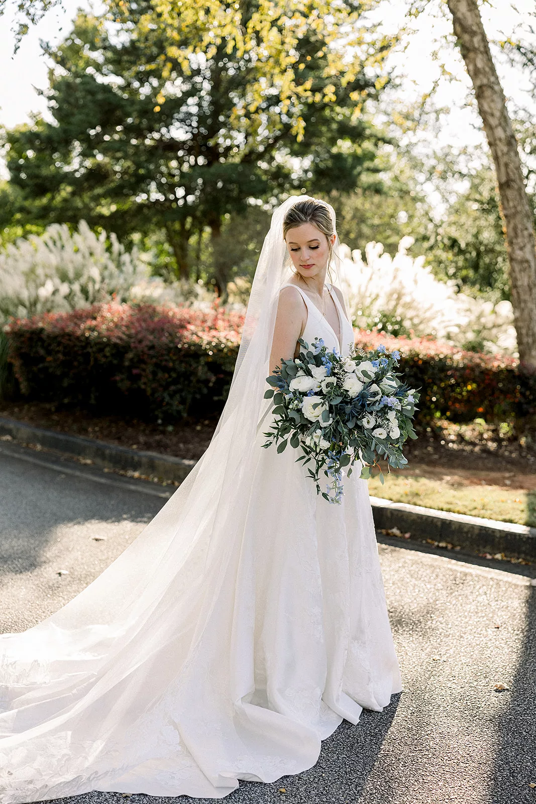 A bride gazes down her shoulder while standing in a garden path while holding her blue and white bouquet