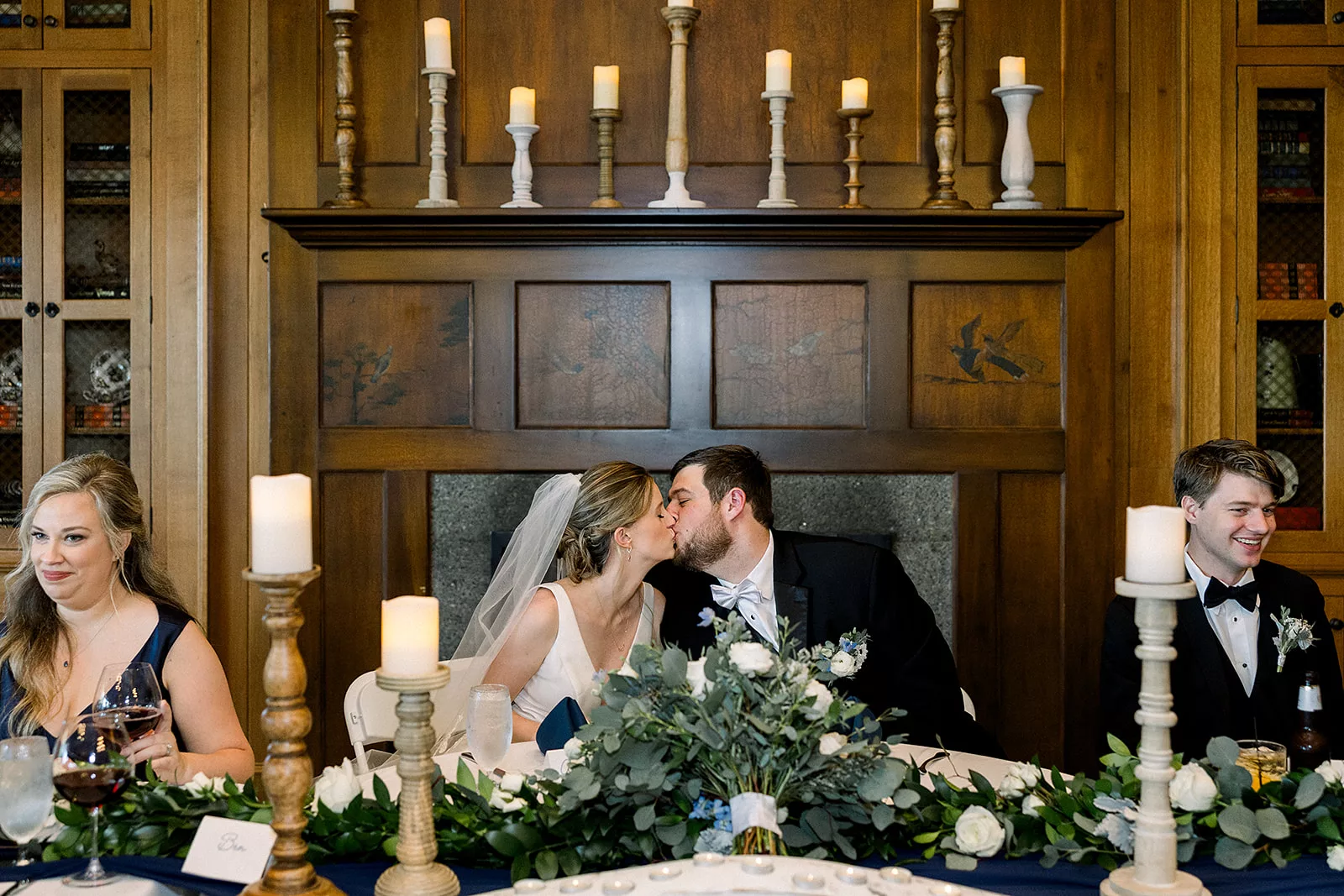 Newlyweds kiss while sitting at their head table in front of a large ornate fireplace at the Curahee Club Wedding venue