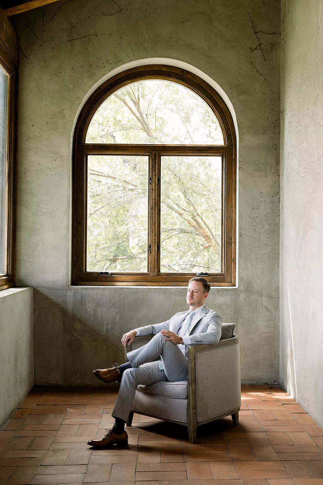 A groom sits in an antique chair in a room with tall round windows