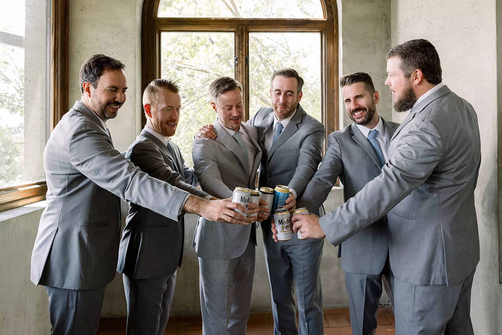 Groomsmen and a groom tap beers together while all wearing grey suits