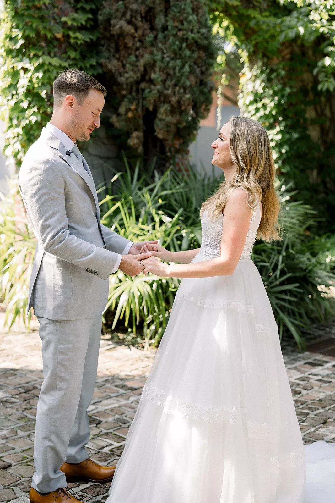 Newlyweds hold hands while standing on a brick patio in a garden covered in ivy