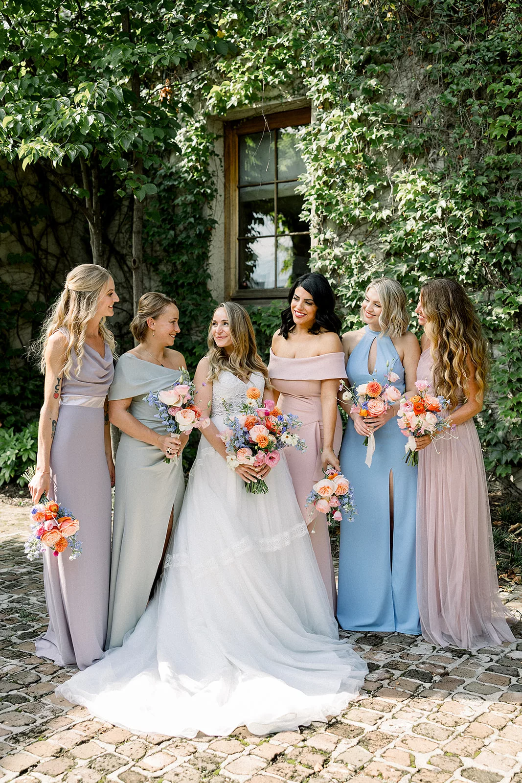 A bride smiles to her bridesmaids as they stand on a patio of an ivy covered building