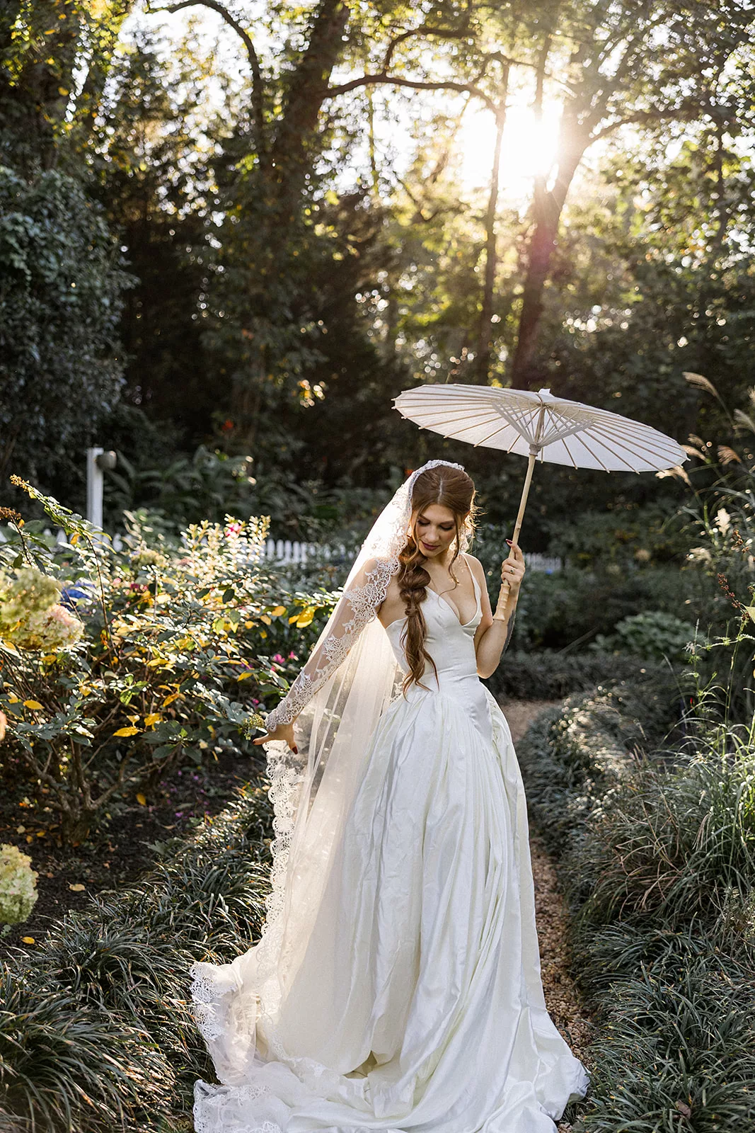 A bride plays with her veil while walking through a Wildflower 301 garden path with an umbrella at sunset