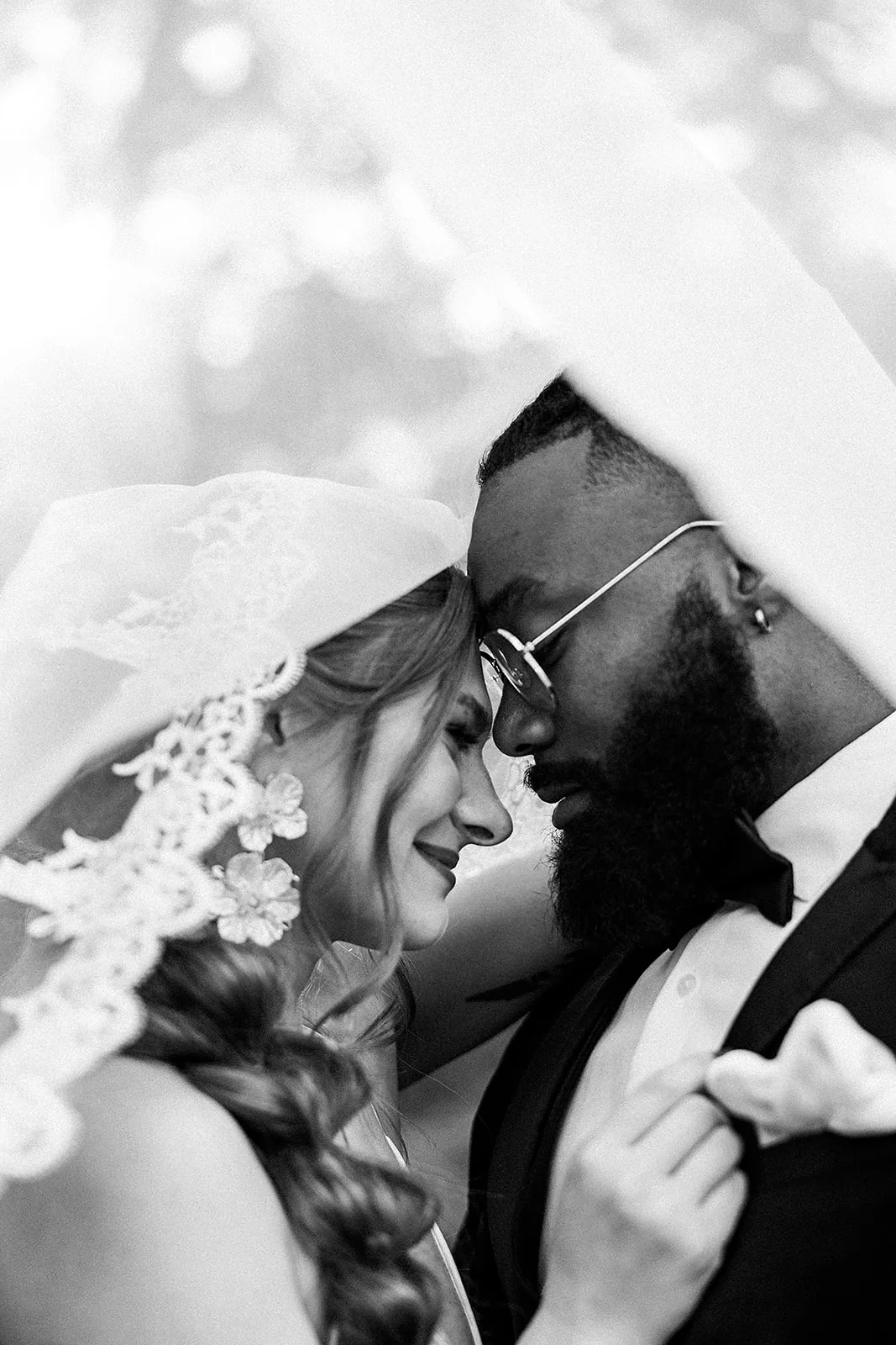 Newlyweds in black and white touch foreheads under a veil in a garden
