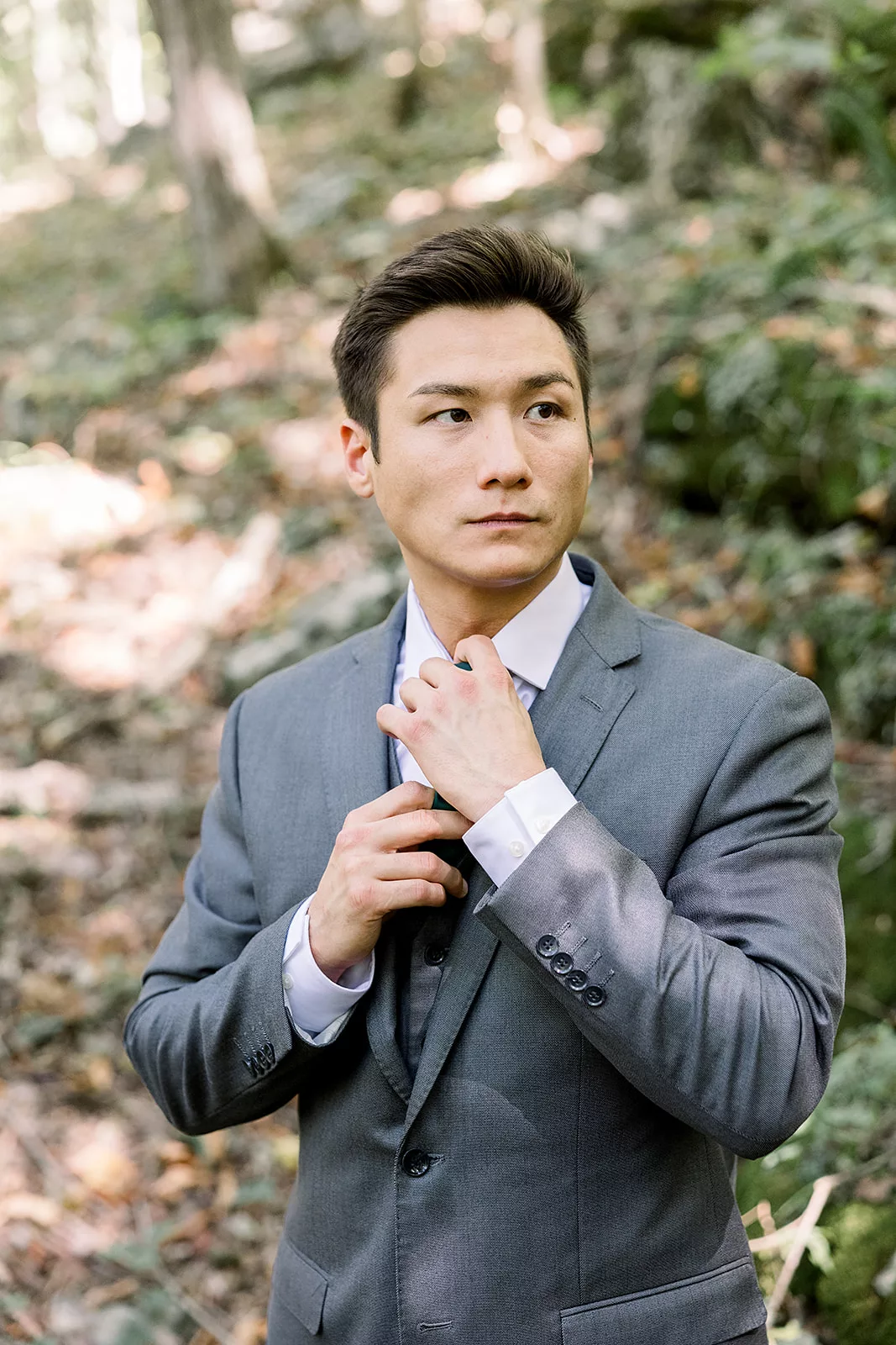 A groom adjusts his tie while standing in a forest in a grey suit