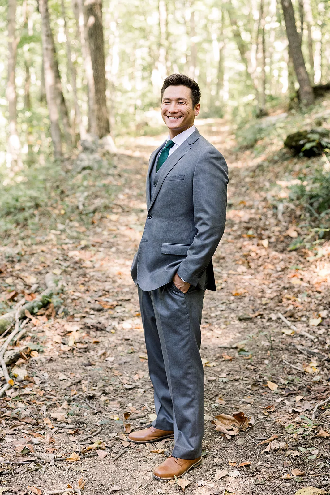 A groom stands in a forest trail with hands in the pockets of his grey suit at a Sustainable Wedding Venue