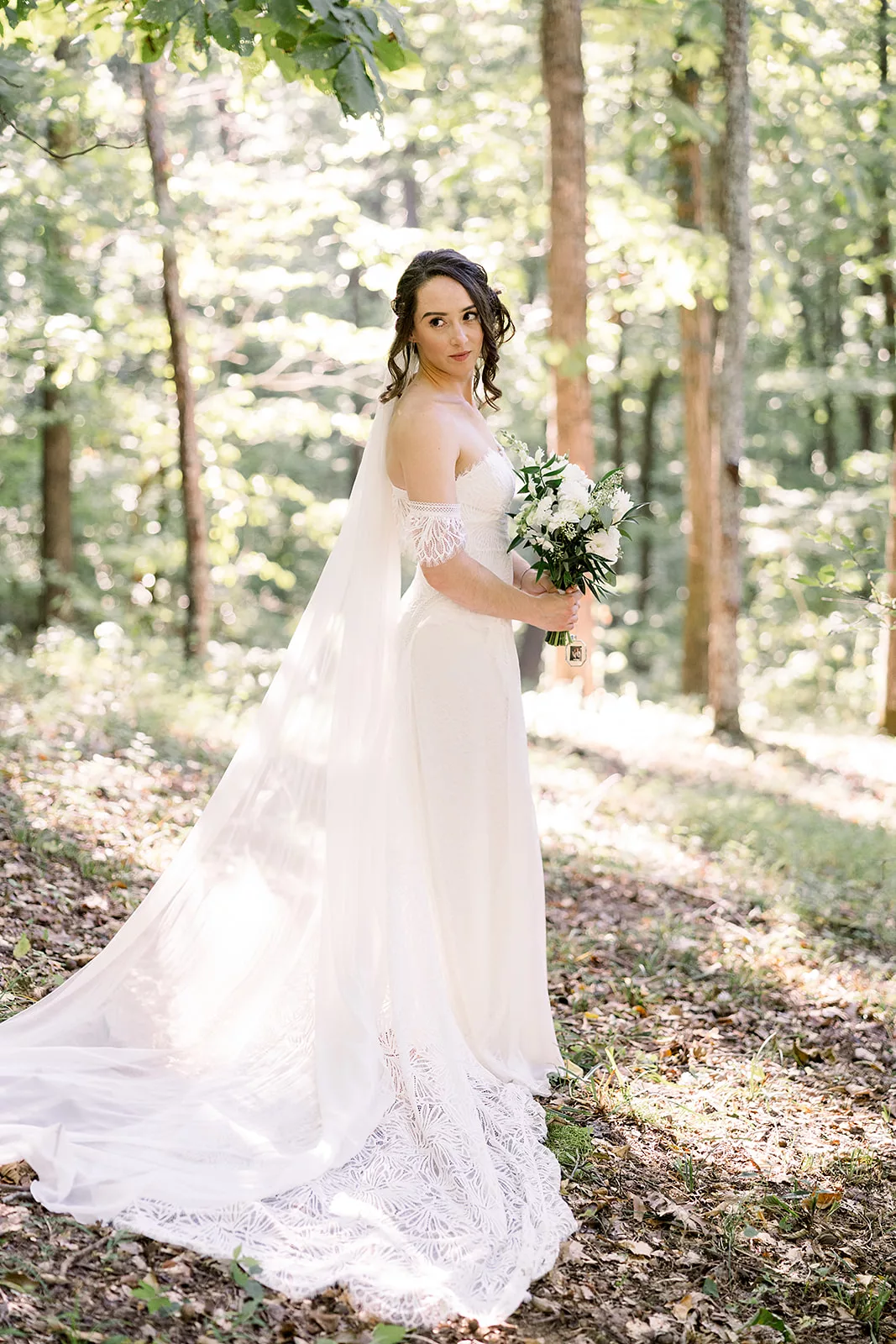 A bride turns to look over her shoulder in a forest while standing and holding her white bouquet Sustainable Wedding Venue