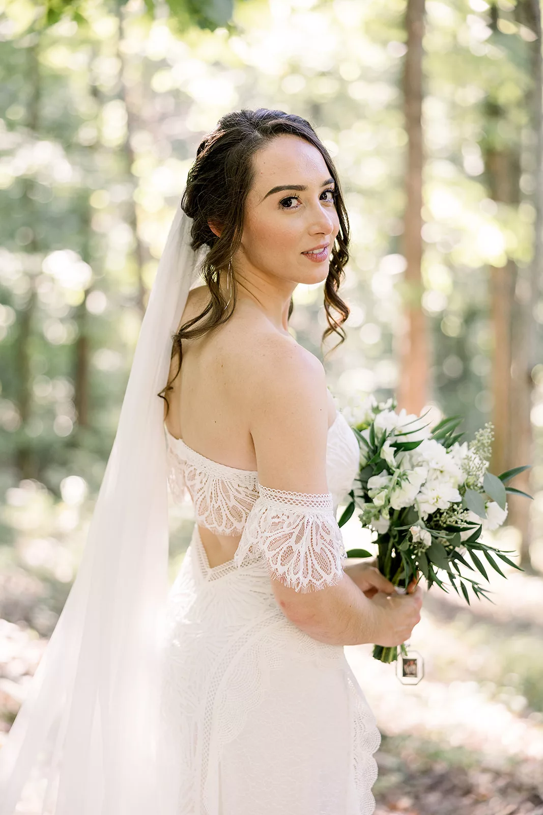 A bride smiles over her shoulder in a lace dress holding a white bouquet in a forest