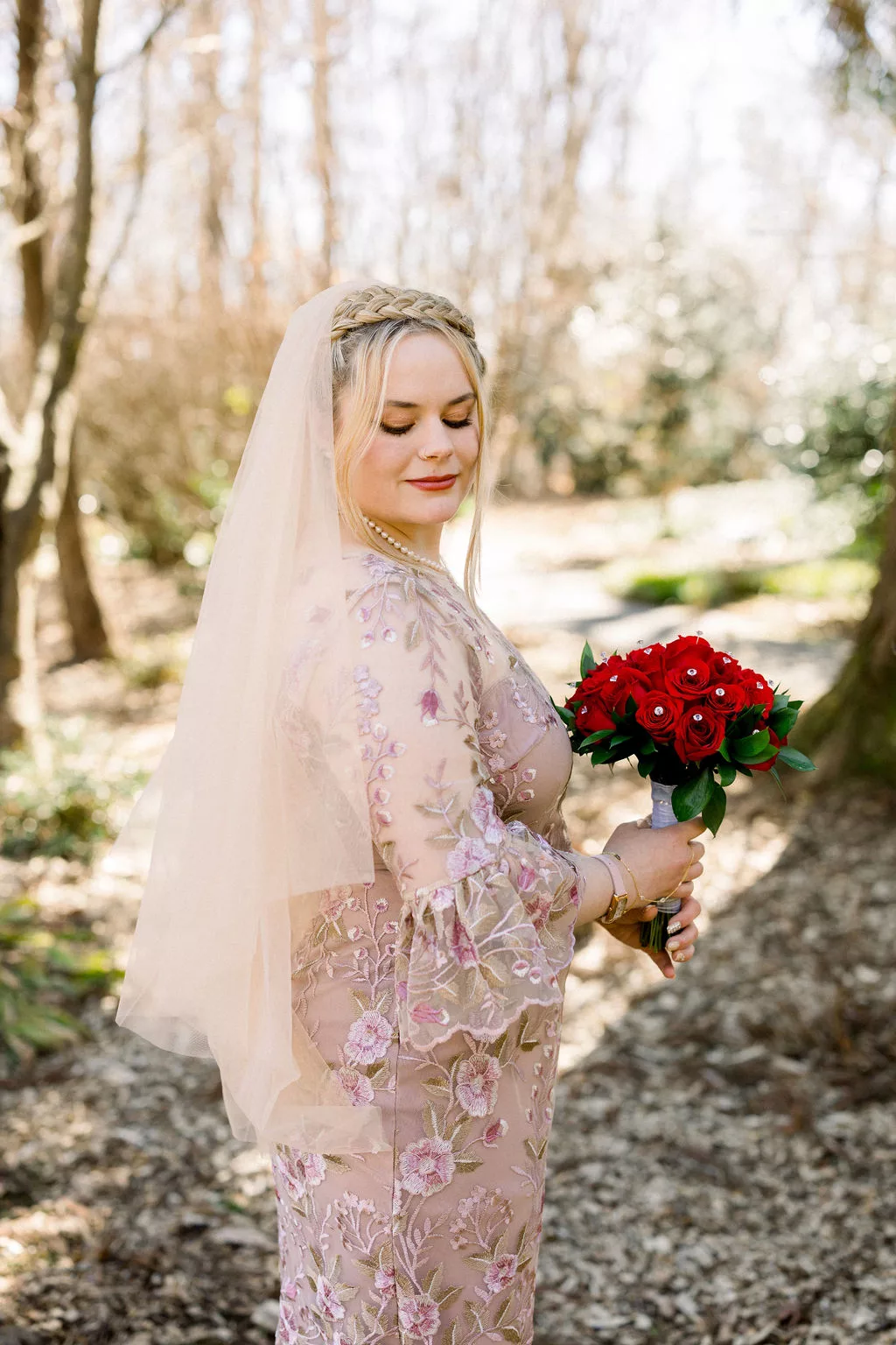 A bride smiles down her shoulder in a pink floral dress while holding a bouquet of red roses at a uga botanical gardens wedding