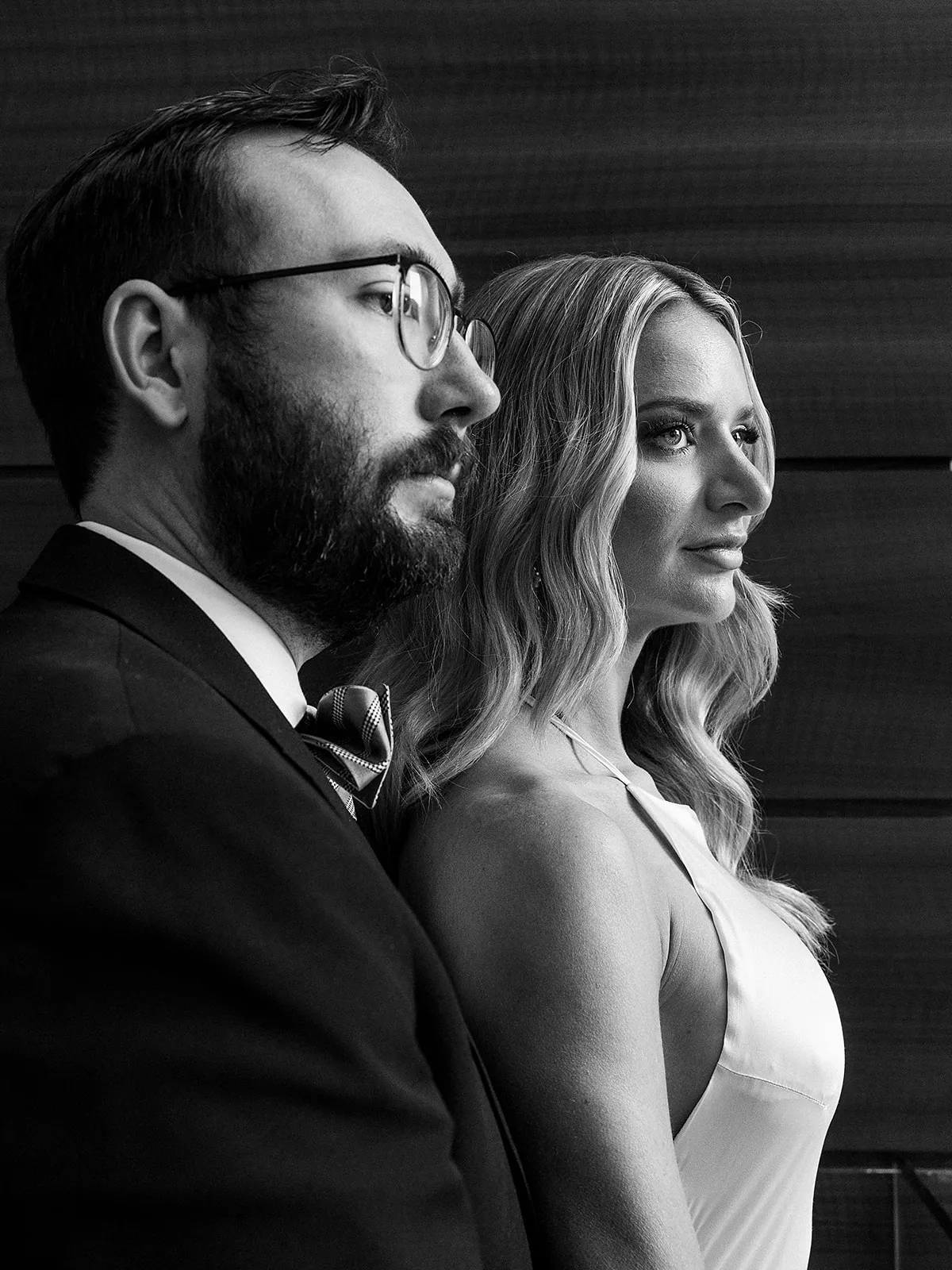 A bride and groom stand together while gazing out a window in black and white